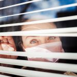 Woman looking out the blind window wearing a surgical mask
